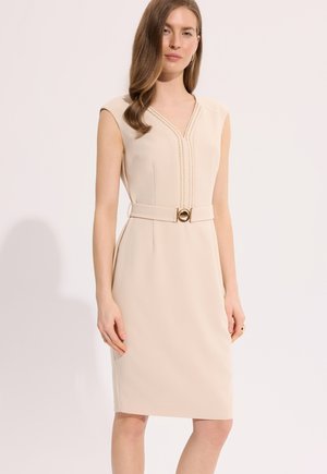 Kobieta w beżowej sukience na ramiączkach z dekoltem w serek, ozdobnym przeszyciem i pasem z dopasowaną złotą okrągłą klamrą, stojąca na białym tle.