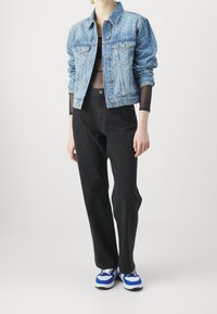 Persona con chaqueta de mezclilla azul con estampado, top negro de manga larga transparente, vaqueros negros y zapatillas azul y blancas, de pie contra un fondo blanco.