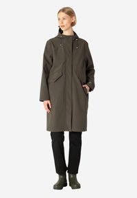 Abrigo impermeable verde oliva con corte recto, capucha ajustable y dos bolsillos frontales. Combinado con pantalones negros y botas verdes.