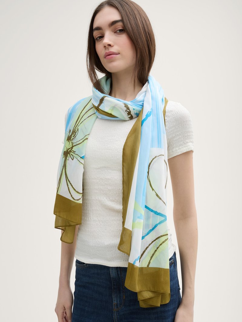 Jeune femme portant un haut blanc texturé à manches courtes, un jean bleu et un foulard en soie aux motifs bleu clair, vert et marron autour du cou.