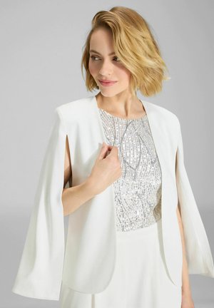 Blonde Frau, die einen weißen Cape-Blazer über einem silbernen, paillettenbesetzten Top trägt, blickt leicht zur Seite mit einem dezenten Lächeln.