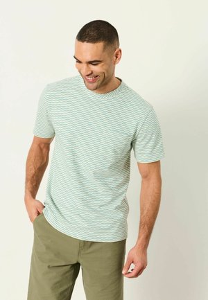 Mann lächelt, trägt ein weißes T-Shirt mit kurzen Ärmeln und blauen wellenförmigen Streifen sowie olivgrüne Hosen, steht vor einem schlichten hellen Hintergrund.