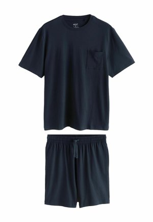Lächelnder Mann mit lockigem Haar, der ein marineblaues T-Shirt und passende Hose trägt, mit den Händen in den Taschen, schaut nach unten vor einem schlichten Hintergrund.