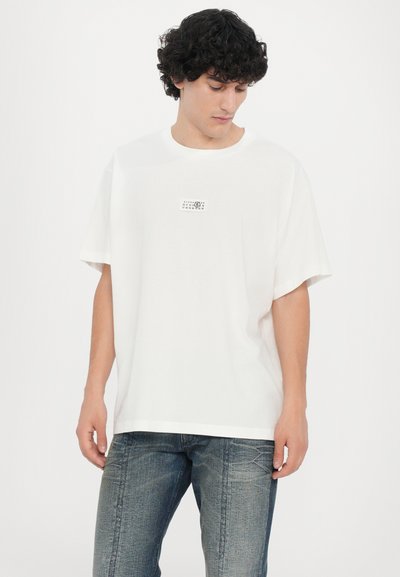 T-shirt en coton blanc à manches courtes et encolure ronde. Présente un petit logo graphique noir sur la poitrine. Porté avec un jean en denim gris.