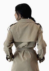 Trench-coat beige avec un col structuré, des épaulettes et une taille ceinturée. Texture lisse avec un dos drapé et des sangles aux poignets.