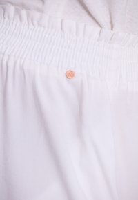 Witte stof met een getextureerde elastische tailleband. Bevat een klein, rond merkteken in lichtperzikkleur. De stof heeft een gladde, zachte textuur.