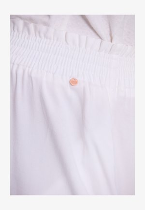 Weißer Stoff mit texturiertem Gummibund. Verfügt über ein kleines, rundes Markenschild in einem hellen Pfirsichton. Der Stoff hat eine glatte, weiche Textur.