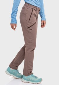 Braune Outdoor-Hosen mit Reißverschlusstaschen, kombiniert mit einem hellblauen Langarmshirt und türkisfarbenen Sportschuhen. Glatter Stoff, taillierte Passform.