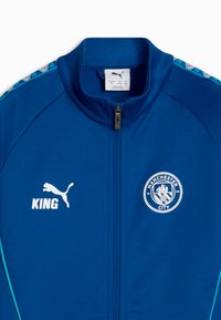 Veste de sport bleue zippée avec le logo de Manchester City sur la poitrine droite, le logo Puma et le texte "KING" sur la poitrine gauche, et des bandes d'épaule blanches et bleues à motif.