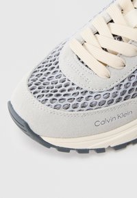 Ανοιχτό γκρι αθλητικό παπούτσι με επάνω μέρος από διχτυωτό υλικό και σουέτ, κρέμ κορδόνια και μαύρη ελαστική σόλα. Διαθέτει λεπτομέρεια με λογότυπο στο πλάι.