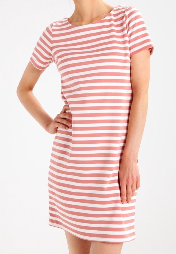 Femme portant une robe courte à manches courtes jusqu'au genou avec des rayures horizontales roses et blanches, une main sur la hanche, sur un fond uni.
