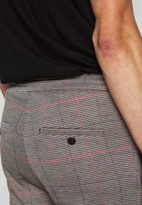 Ruudulliset housut mustassa, valkoisessa ja punaisessa värissä, valmistettu teksturoidusta kankaasta. Takana on tasku mustalla nappidetaililla.