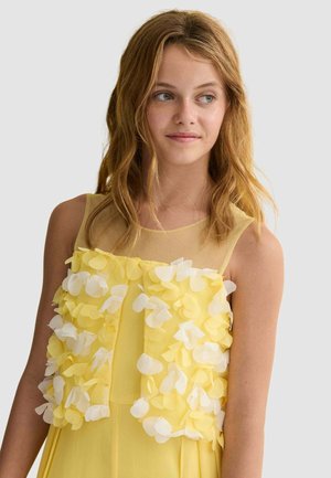 Ragazza giovane con capelli biondi mossi che indossa un vestito giallo senza maniche decorato con applicazioni a forma di petali bianchi e gialli.