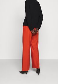 Schwarzer gerippter Pullover, rote weite Hose und schwarze spitze High Heels. Die Figur steht mit hinter dem Rücken verschränkten Händen und ist von der Kamera abgewandt.