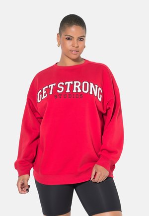 Punainen ylisuuri huppari, jossa on valkoisella ja mustalla tulostettu "GET STRONG STUDIOS", pyöreä kaula-aukko ja pitkät hihat.