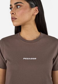Smeđa pamučna majica kratkih rukava s bijelim logom "PEGADOR" na prsima. Jednostavan, prilagođen oblik s okruglim izrezom.
