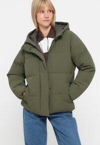 Chaqueta acolchada verde con capucha, que cuenta con un cierre de botones en el frente, bolsillos laterales y un diseño acolchado. Lleva encima de una blusa de capas marrón y blanca.