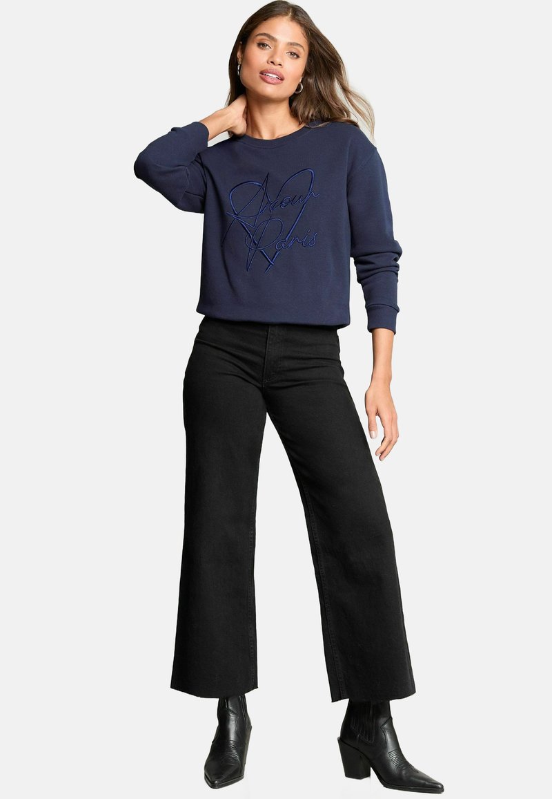 Felpa blu navy con ricamo "Amour Paris", abbinata a jeans neri a vita alta con gamba larga e stivaletti neri con tacco.