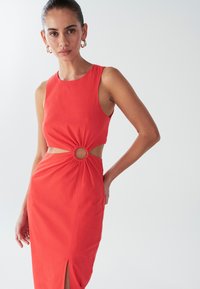 Crvena haljina bez rukava s okruglim izrezom u struku, bočnim prorezom i naboranim detaljima, izrađena od glatkog, laganog materijala.