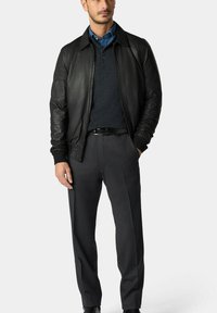 Černá kožená bomber bunda s žebrovanými manžetami, nošená přes tmavě modrý svetr a košili s límečkem, zkombinovaná se šedými oblekovými kalhotami.