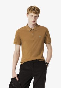 Camisa polo de algodón marrón con una tapeta de botones y un pequeño logo de Lacoste. Modelo vistiendo pantalones oscuros de pana, sosteniendo una chaqueta negra.