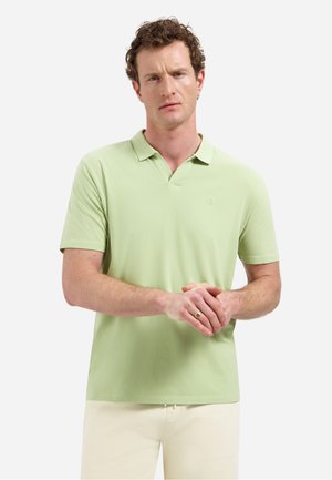 Man met krullend haar die een lichtgroen poloshirt met korte mouwen en crèmekleurige broek draagt, staand met gevouwen handen tegen een witte achtergrond.