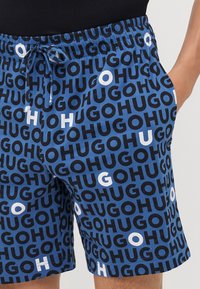 Shorts bleus avec un motif logo noir et blanc répété, taille élastique avec cordon de serrage et poches latérales. Matière en coton.