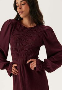 Robe bordeaux avec des manches bouffantes et un détail de smocks texturés au niveau du corsage. Les manches longues présentent des poignets froncés pour un look volumineux.