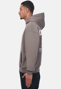 Hellgrauer Kapuzenpullover mit Kängurutasche, oversized Schnitt und weißem Textdruck. Rippbündchen und -saum. Der Stoff wirkt weich und langlebig.