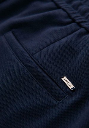 Närbild av marinblått texturerat tyg med en sydd ficka och en liten metalltagg märkt "ombre" som är fäst nära fickan.