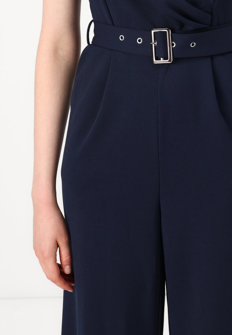 Tuta navy con un design strutturato, dotata di una cintura larga con una fibbia rettangolare in argento e grommet decorativi. Tessuto con una consistenza liscia.