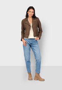 Hnědá surová kožená motorkářská bunda se zipy, nošená přes béžový top. V kombinaci se světlými modrými džíny a světle hnědými šněrovacími botami s robustními podrážkami.