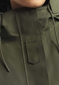 Veste vert olive avec un col montant, une poche kangourou sur la poitrine et des cordons de serrage. Texture lisse avec des détails de couture subtils.