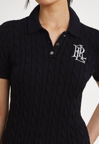 Pletené polo tričko v námorníckej modrej farbe s vrkočovým vzorom, golierom a dvoma gombíkmi. Na hrudi má vyšitú bielu značku.