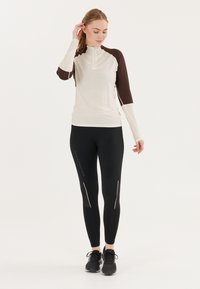 Langærmet, lynlås athletic top i crème med brune detaljer, parret med sorte leggings med reflekterende striber og logo, båret med sneakers.