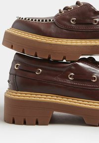 Ruske nahkate kengät paksulla ruskealla pohjalla, koristeelliset ompeleet ja metalliset nyörit. Ominaisuuksiin kuuluu kerrostettu muotoilu ja teksturoidut pinnat.