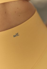 Keltainen urheilullinen lyhyt toppi, jossa on sileä pinta. Helmassa sininen "self."-logo, saumaton muotoilu mukavuutta varten.
