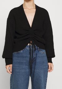 Svart stickad tröja med djup V-hals, rynkad framsida och dragsko i fållen. Har överdimensionerade ärmar och en texturerad tyg. Parat med blå jeans.