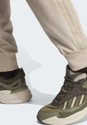 Osoba ubrana w beżowe spodnie dresowe i oliwkowozielone tenisówki z białymi podeszwami, stojąca na jasnej nawierzchni.