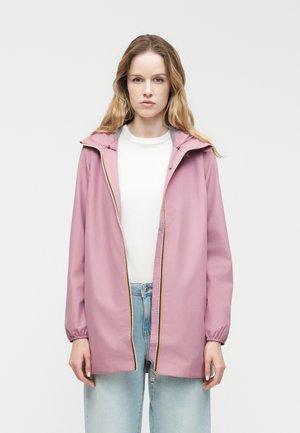 Kvinde med langt blondt hår iført en lyse lilla hættetrøje over en hvid skjorte og lyseblå jeans, stående mod en ensfarvet baggrund.