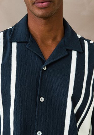 Mees, kes kannab tumel sinist kaelusega särki, millel on vertikaalsed valged triibud ja nähtavad nööbid, beež taust.