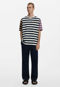 Short-sleeve striped t-shirt in navy, white, and mustard with a relaxed fit; paired with dark denim pants and black slide sandals.