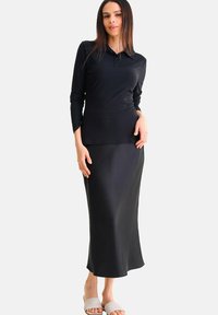 Zwarte polo met lange mouwen en ribbelstructuur gecombineerd met een gladde zwarte maxi-rok. Het model draagt witte slidesandalen.