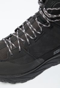 Schwarzer Wanderschuh mit strukturiertem Obermaterial, reflektierenden Schnürsenkeln und einer dicken, robusten Sohle mit geometrischen Mustern für besseren Halt.