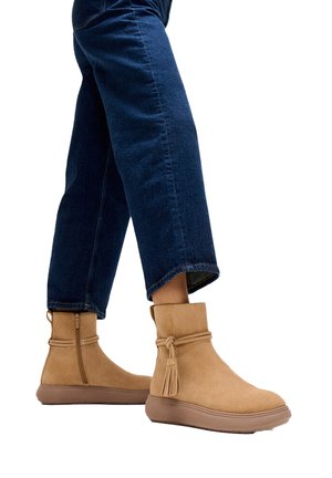 Persoon die een blauwe spijkerbroek en tan enkellaarzen met zijrits en decoratieve kwast draagt tegen een witte achtergrond.