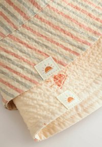 Gequilte deken met afwisselende strepen in roze, blauw en crème. Opvallende zon grafische labels en een zachte beige onderkant met textuur.