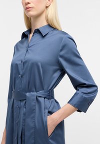 Blaues Satinhemdkleid mit Kragen, langen Ärmeln, Taillenbindegürtel, vorderen Knöpfen und seitlichen Taschen. Glatte Textur über das gesamte Kleid.