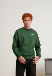 Sudadera de algodón verde con cuello redondo y puños acanalados, que presenta un pequeño logo blanco de Nike en el lado izquierdo del pecho. Se lleva con pantalones negros.