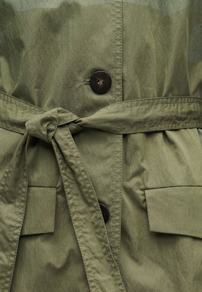 Ελαφρύ μπουφάν σε ελιά πράσινο με ζωνάκι στη μέση που δένει, κούμπωμα με κουμπιά και δύο μπροστινές τσέπες. Ομαλή υφή με ορατές ραφές.