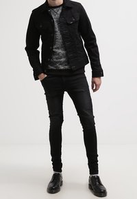 Giacca in denim nera con due tasche sul petto, maglione grigio a fantasia, jeans neri slim-fit e scarpe nere con lacci. Look casual.